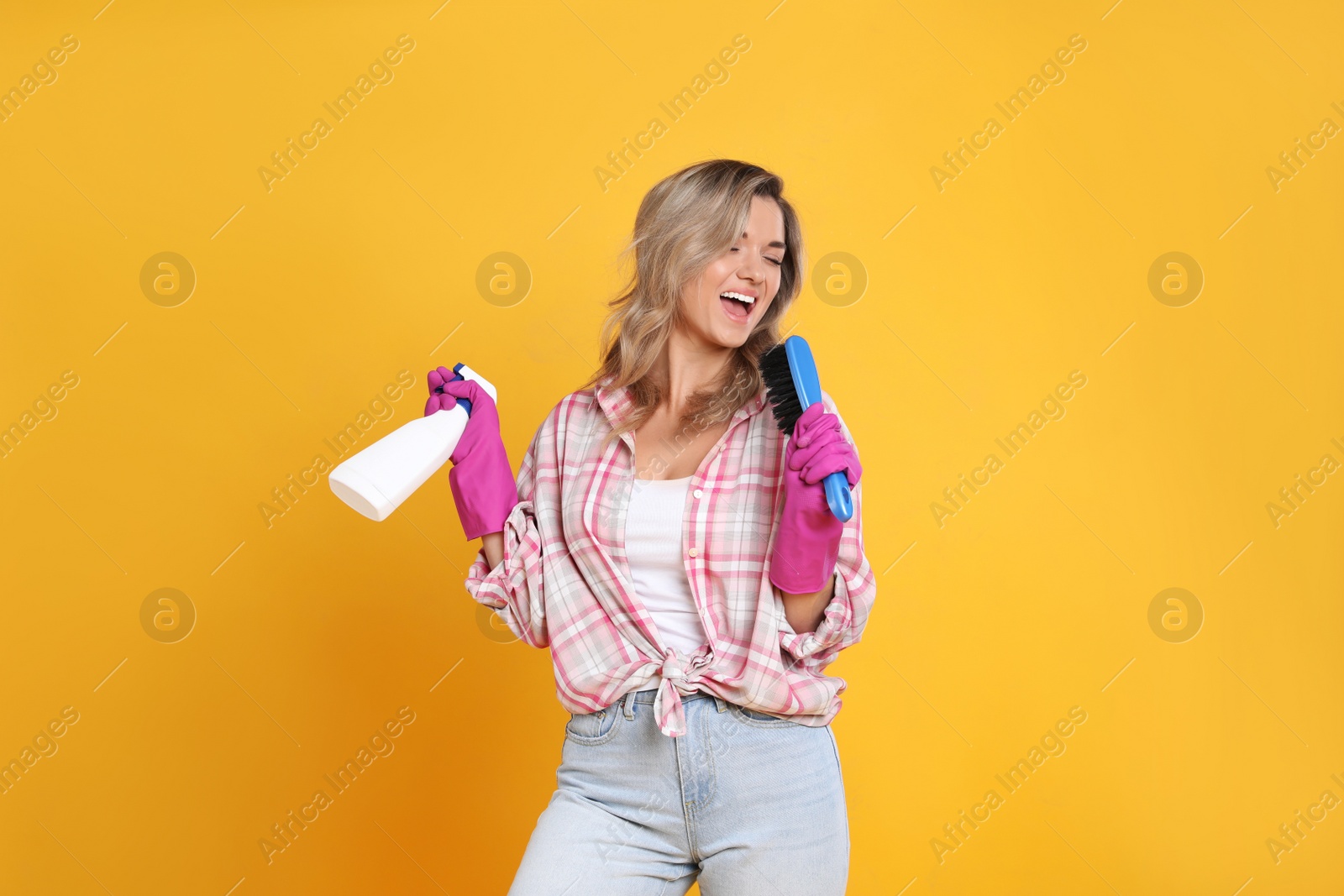Photo of Beautiful young woman with brush and bottle of detergent singing on orange background
