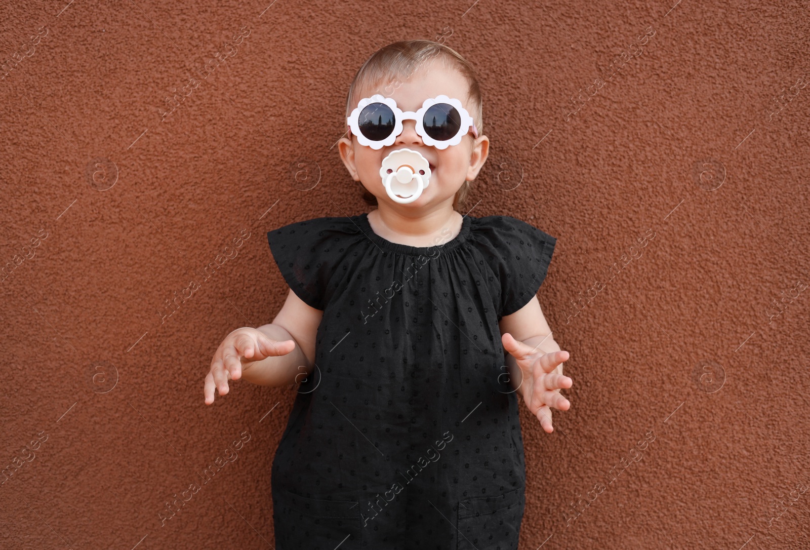 Photo of Cute little girl wearing stylish clothes with sunglasses and pacifier near brown wall