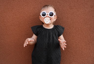 Photo of Cute little girl wearing stylish clothes with sunglasses and pacifier near brown wall