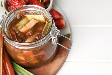 Photo of Jar of tasty rhubarb jam, fresh stems and strawberries on white wooden table, closeup. Space for text