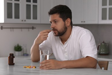 Depressed man with glass of water taking antidepressant pill at table in kitchen