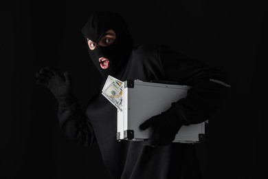 Emotional thief in balaclava with briefcase of money on black background
