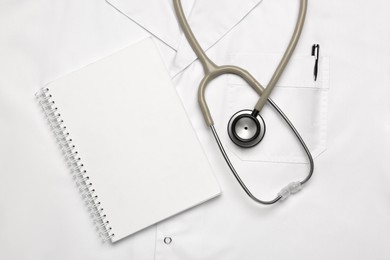 Stethoscope and notepad on white medical uniform, top view