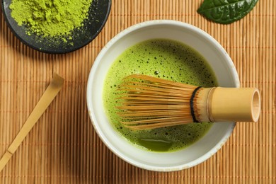 Photo of Cup of fresh matcha tea with whisk, spoon and green powder on bamboo mat, flat lay