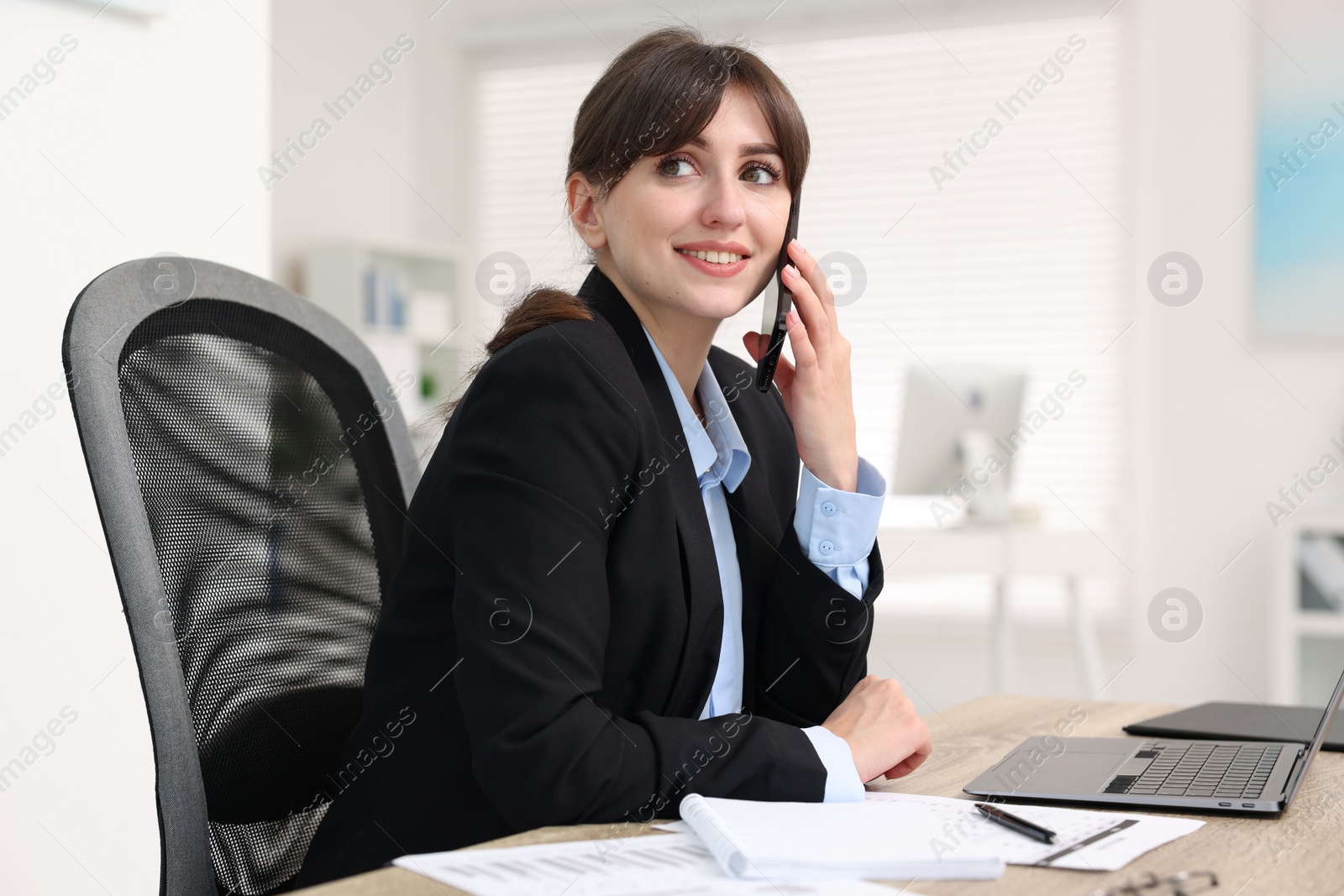 Photo of Smiling secretary talking by smartphone at table in office