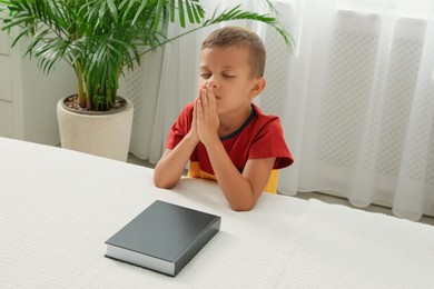 Cute little boy with hands clasped together saying prayer over Bible at home