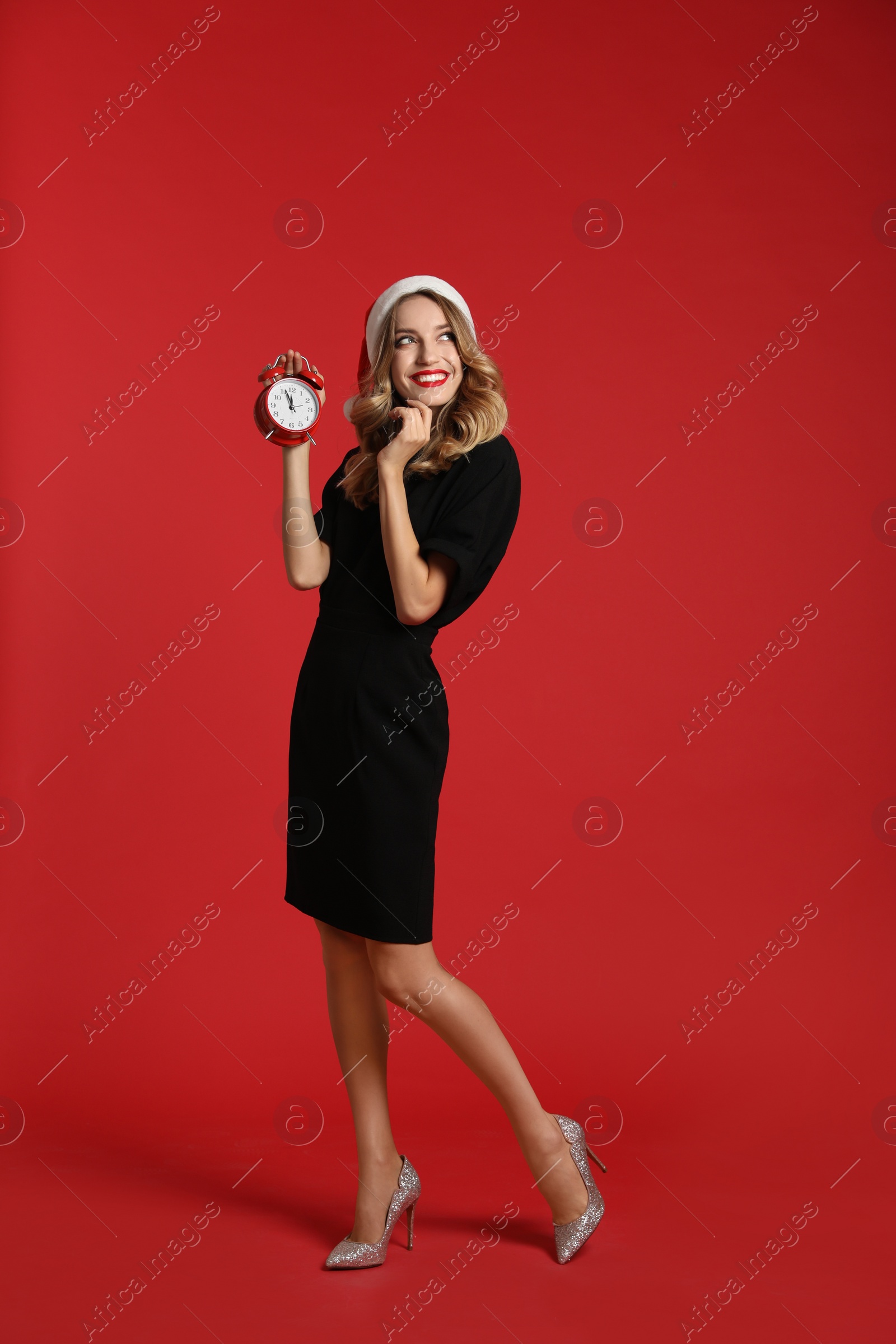 Photo of Happy young woman wearing Santa hat with alarm clock on red background. Christmas time