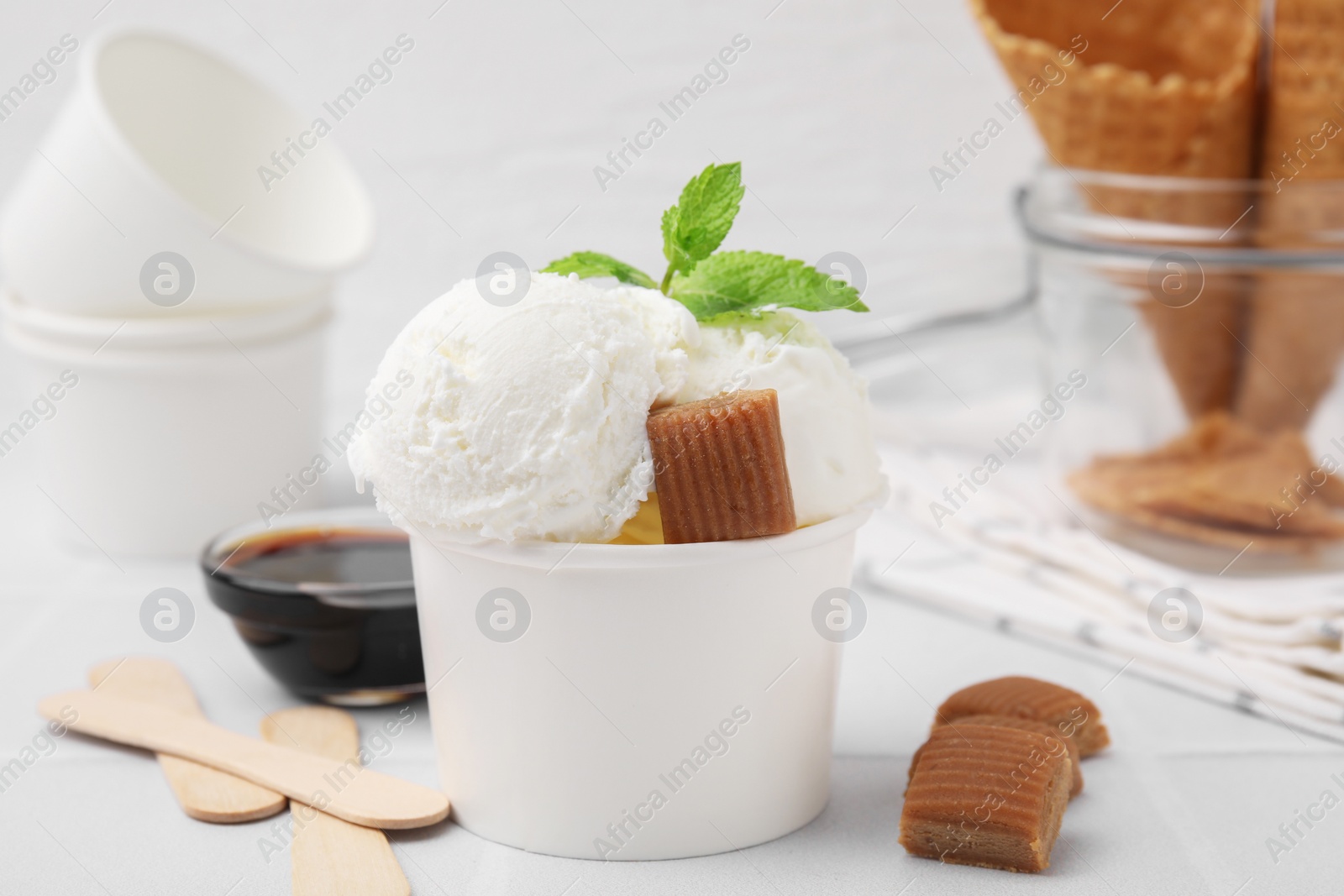Photo of Scoops of tasty ice cream with caramel candies, sauce and mint leaves on white tiled table, closeup
