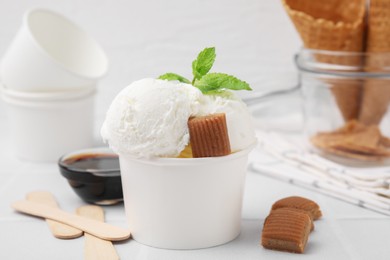 Scoops of tasty ice cream with caramel candies, sauce and mint leaves on white tiled table, closeup