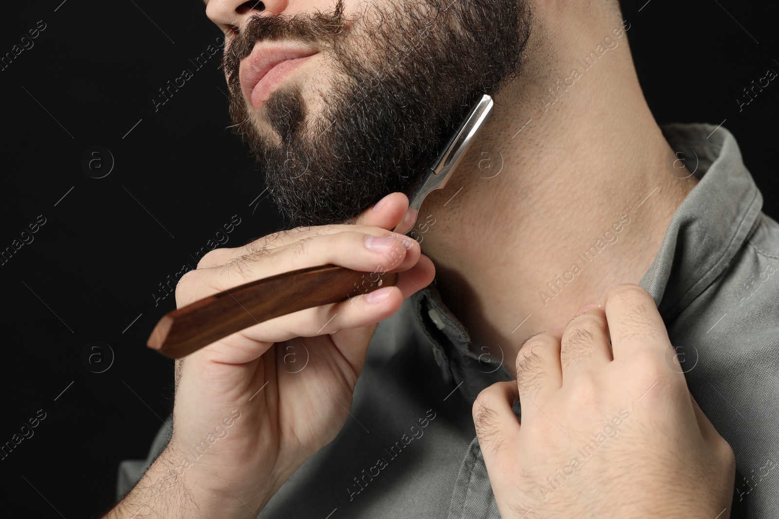 Photo of Handsome young man shaving beard with blade on black background, closeup