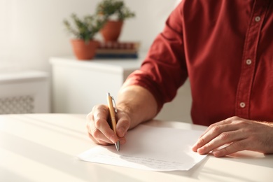 Photo of Man writing letter at white table in room, closeup