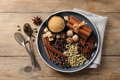 Plate with different aromatic spices and spoons on wooden table, flat lay