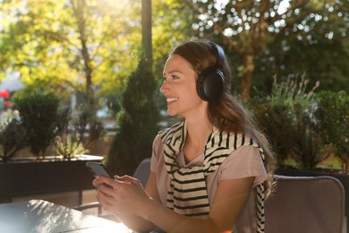 Photo of Smiling woman in headphones using smartphone in outdoor cafe. Space for text
