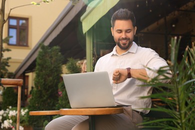 Photo of Handsome young man with laptop checking time on watch at table in outdoor cafe. Space for text