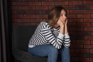 Photo of Sad young woman sitting on chair near brick wall indoors