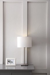 Photo of Wooden chest of drawers with picture and lamp near white wall in room