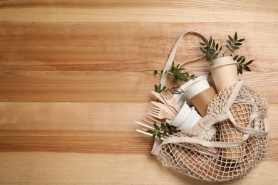 Photo of Net bag with disposable tableware and green twigs on wooden background, top view. Space for text