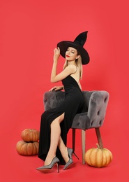 Photo of Beautiful woman wearing witch costume in armchair and pumpkins on red background. Halloween party