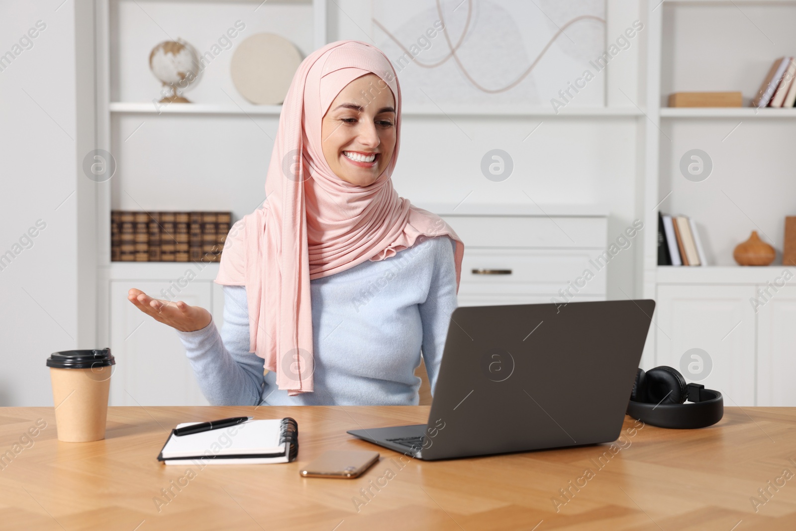 Photo of Muslim woman in hijab using video chat on laptop at wooden table in room