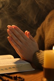 Woman praying at table with burning candle and Bible, closeup