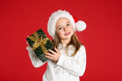 Photo of Cute child in Santa hat with Christmas gift on red background
