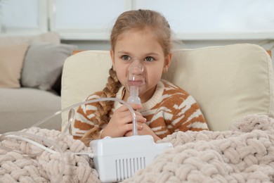 Photo of Little girl using nebulizer for inhalation in armchair at home
