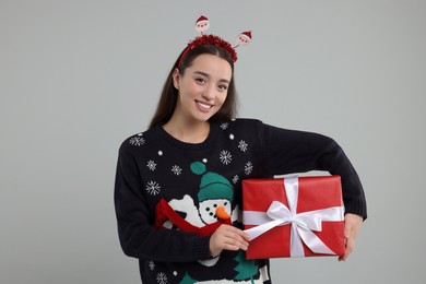 Happy young woman in Christmas sweater and Santa headband holding gift on grey background