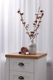 Photo of Alarm clock and vase with cotton branches on wooden commode indoors