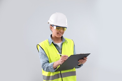 Photo of Female industrial engineer in uniform with clipboard on light background. Safety equipment