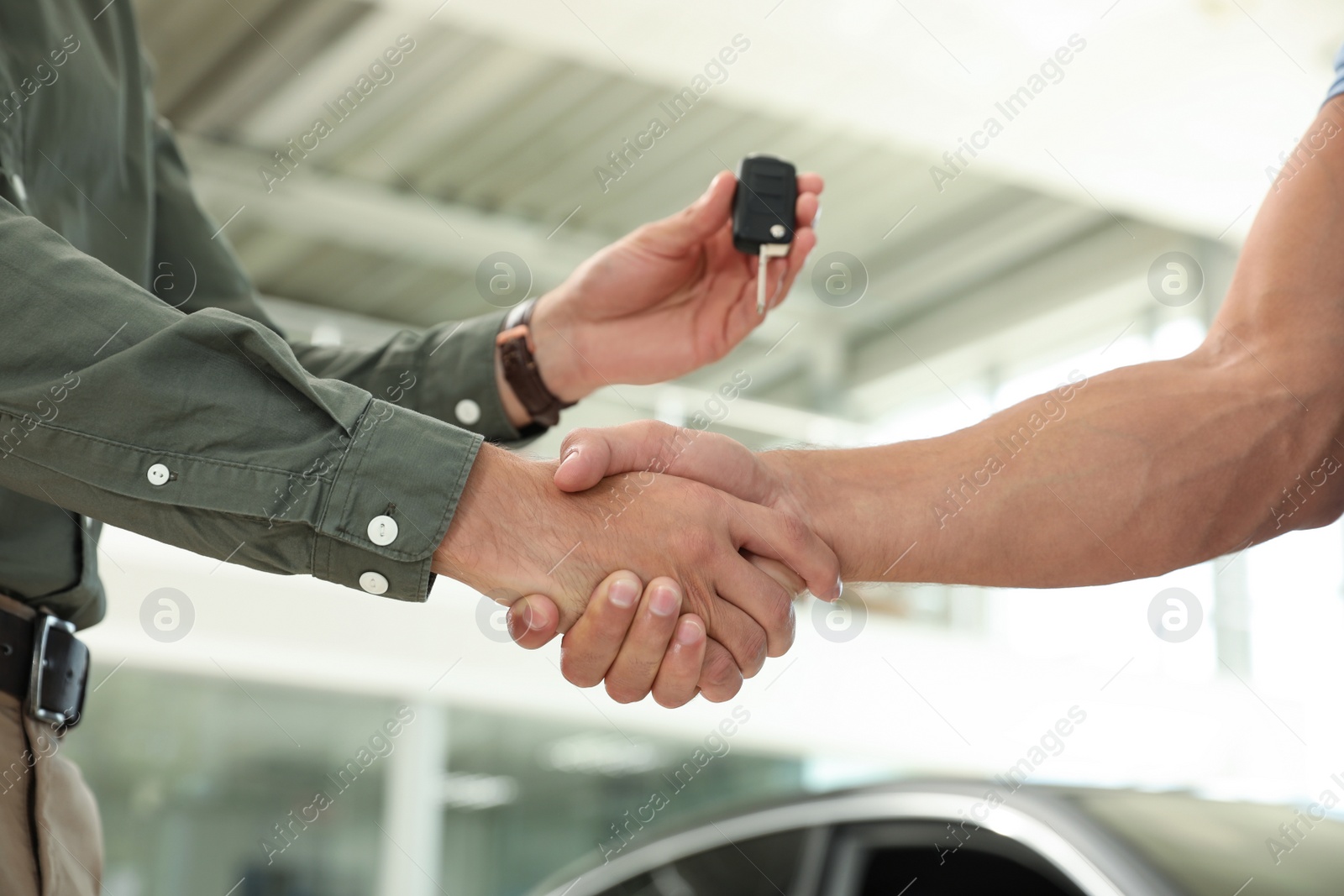 Photo of Young salesman shaking hands with customer and giving car key in modern auto dealership, closeup