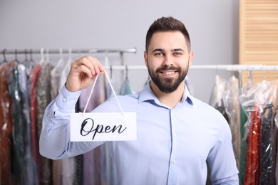 Photo of Dry-cleaning service. Happy worker holding Open sign indoors