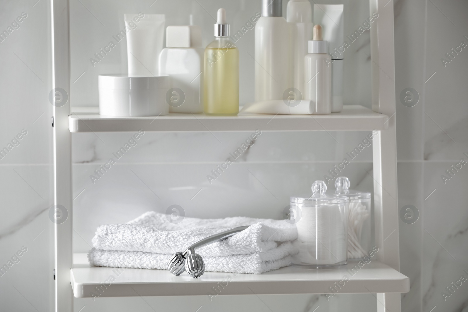 Photo of Metal face roller, towels and cosmetic products on shelving unit in bathroom