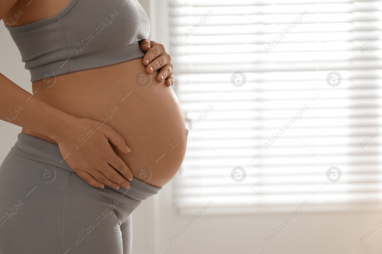 Photo of Pregnant young woman touching belly near window indoors, closeup. Space for text