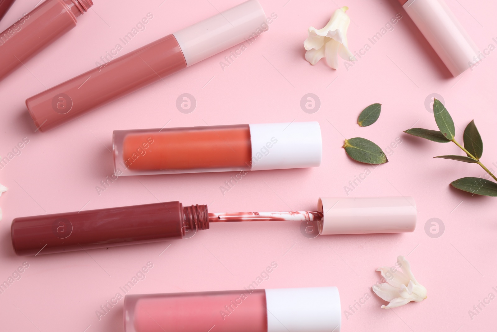 Photo of Different lip glosses, applicator, flowers and green leaves on pink background, flat lay