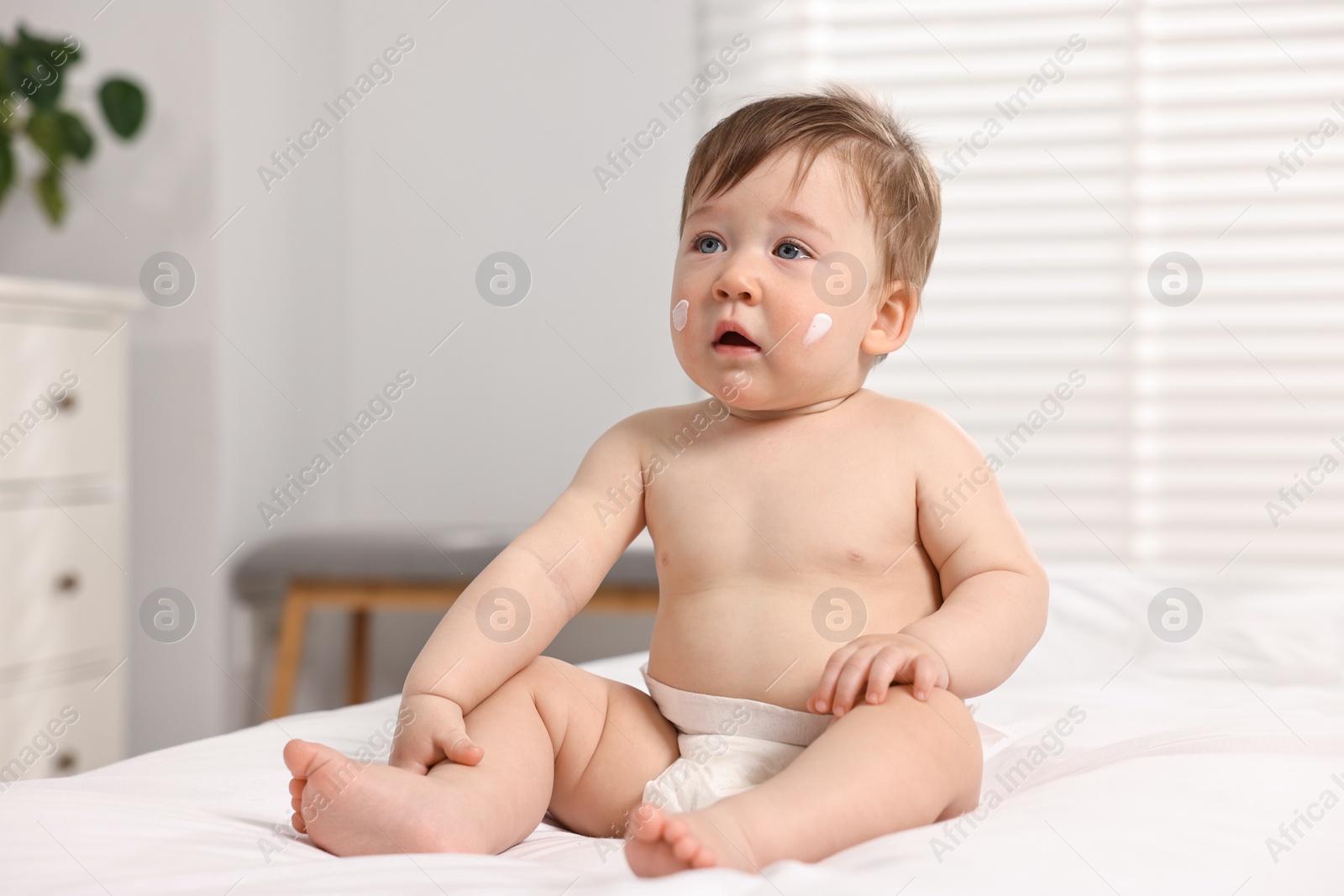 Photo of Cute little baby with moisturizing cream on face sitting on bed indoors