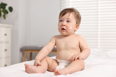 Cute little baby with moisturizing cream on face sitting on bed indoors