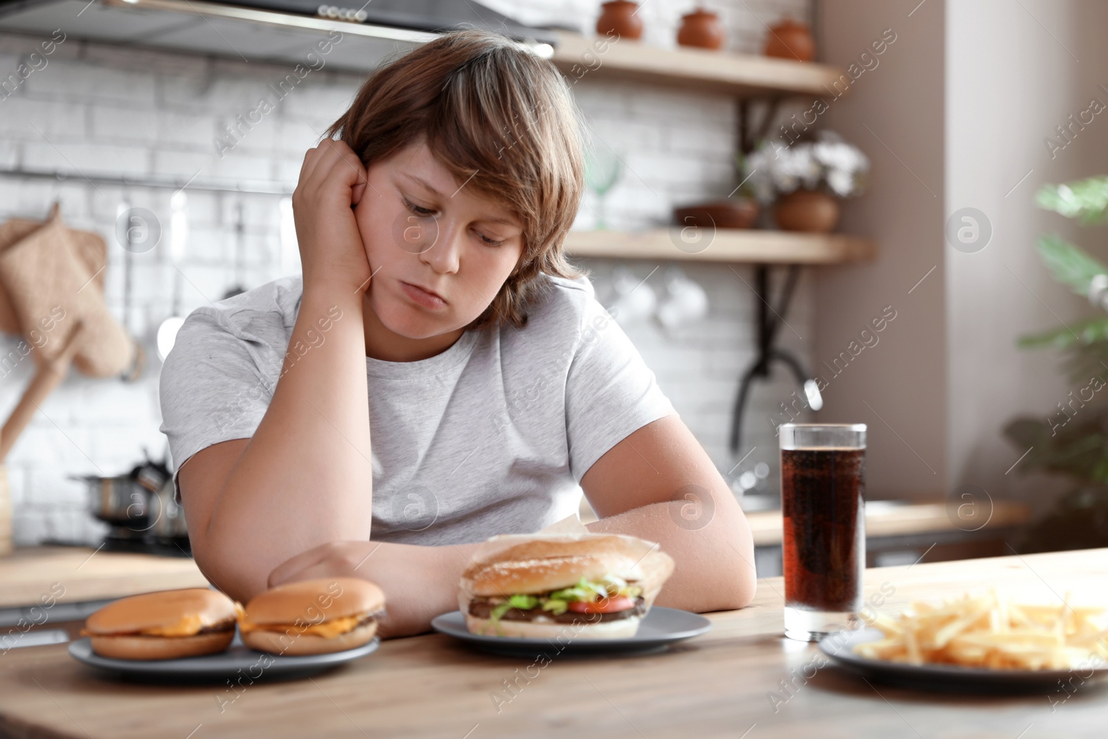 Photo of Emotional overweight boy at table with fast food in kitchen