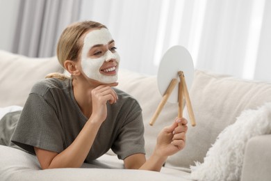 Photo of Young woman with face mask looking into mirror on sofa at home. Spa treatments