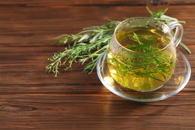 Photo of Cup of homemade herbal tea and fresh tarragon leaves on wooden table. Space for text