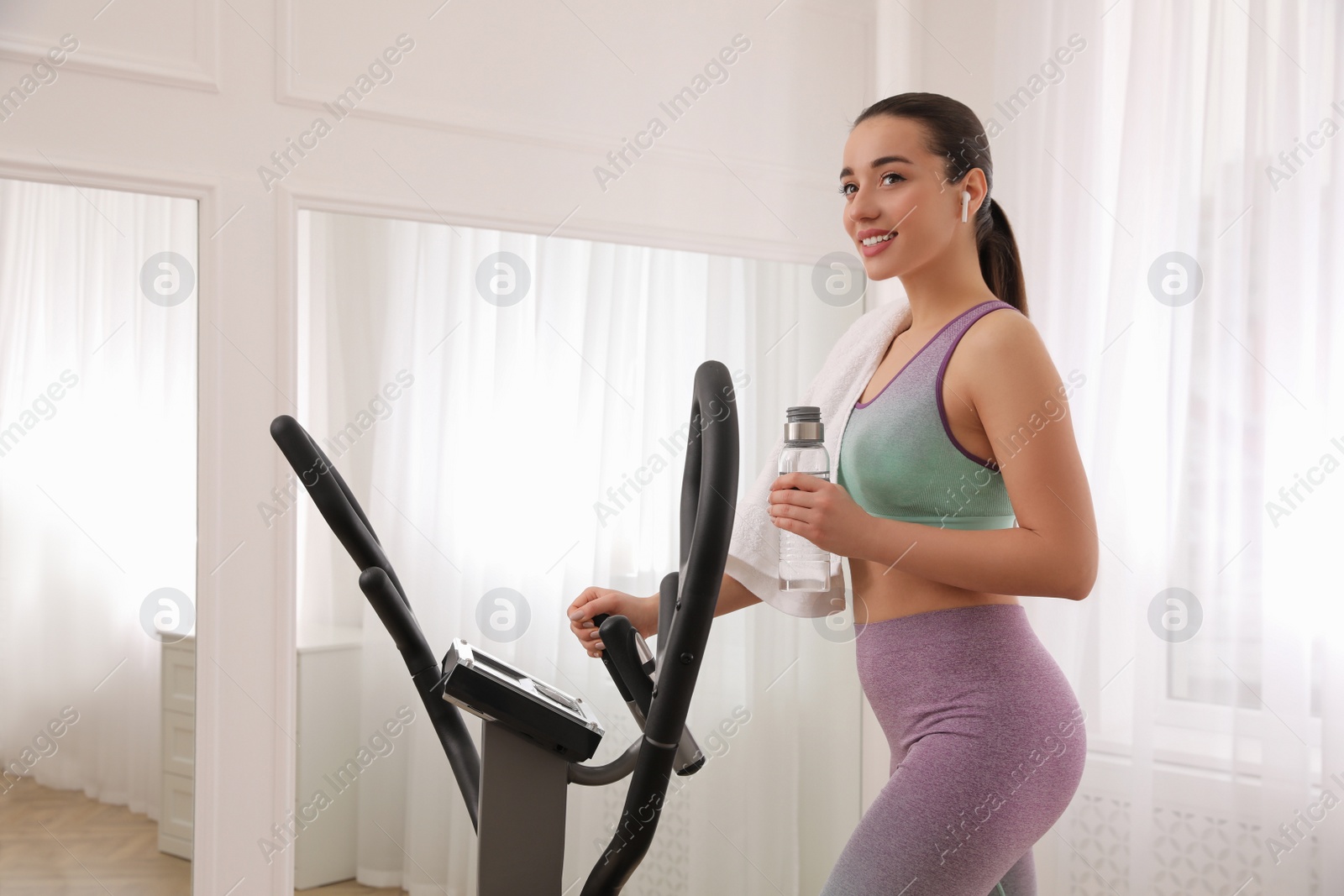 Photo of Woman with bottle and towel using modern elliptical machine at home