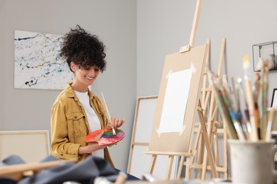 Photo of Young woman mixing paints on palette with brush near easel in studio