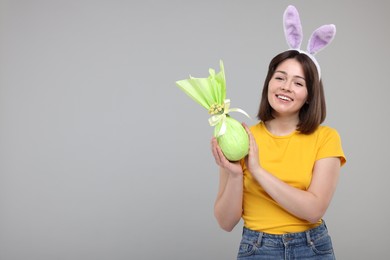 Photo of Easter celebration. Happy woman with bunny ears and wrapped egg on grey background, space for text