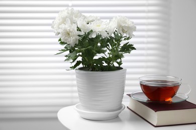 Beautiful chrysanthemum plant in flower pot, cup of tea and book on white table indoors, space for text