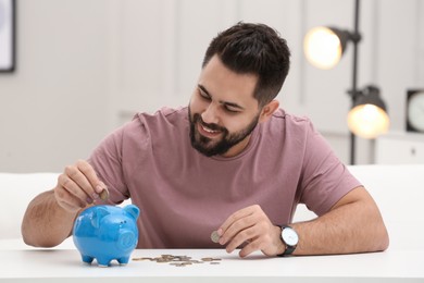 Happy young man putting money into piggy bank at white table indoors