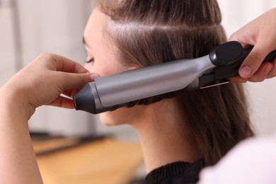 Hairdresser curling woman's hair with iron in salon, selective focus