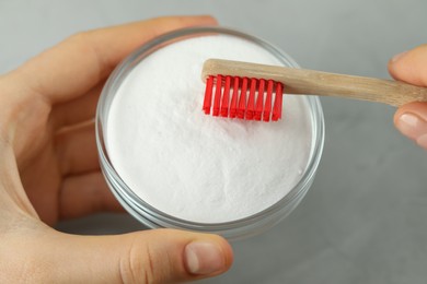 Woman with toothbrush and bowl of baking soda at grey table, closeup