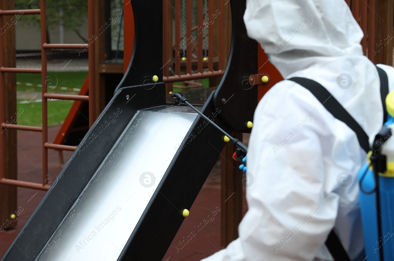 Photo of Woman wearing chemical protective suit at playground, focus on disinfectant sprayer. Preventive measure during coronavirus pandemic