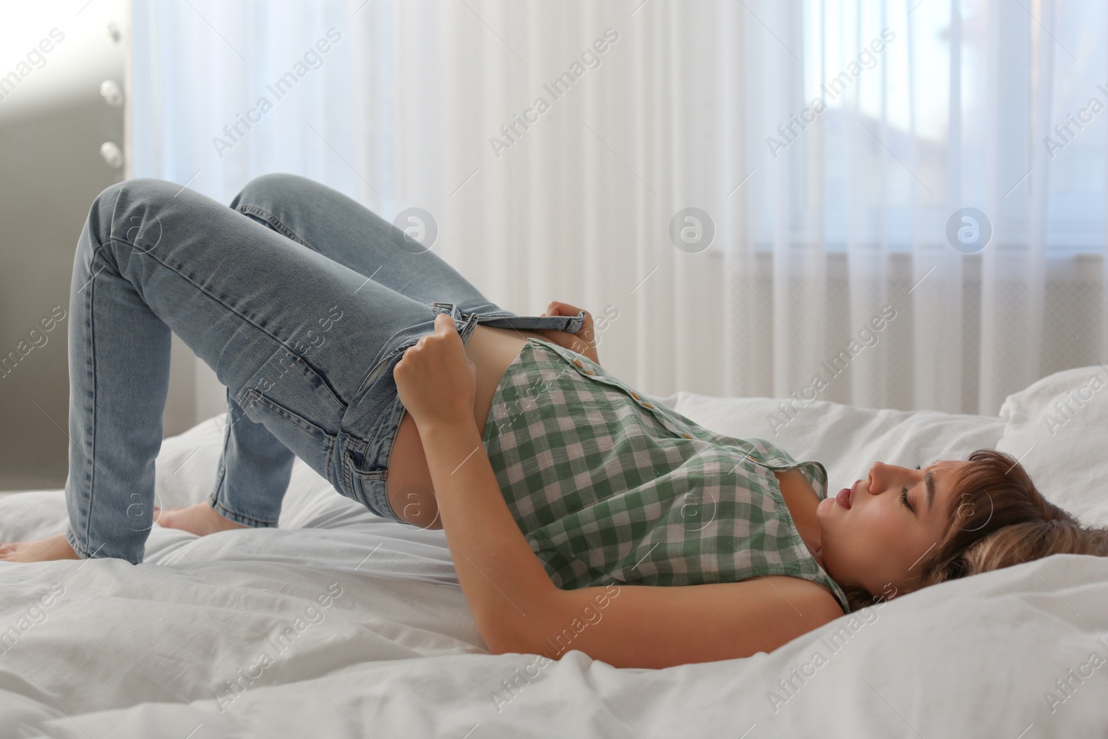 Photo of Young woman struggling to squeeze into tight jeans while lying on bed at home