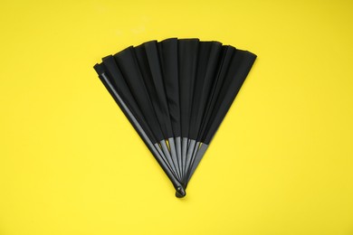 Stylish black hand fan on yellow background, top view