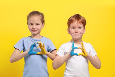 Photo of Little girl and boy making heart with their hands painted in Ukrainian flag colors on yellow background. Love Ukraine concept